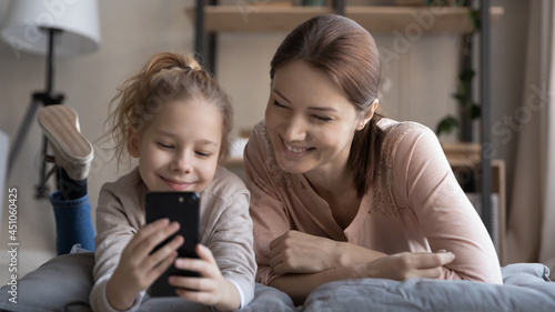 Happy young mom and small teen daughter relax together have webcam online digital virtual event on smartphone. Smiling mother and little girl child talk speak on video call on cellphone.