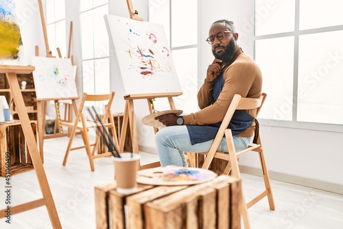 African american artist man painting on canvas at art studio serious face thinking about question with hand on chin, thoughtful about confusing idea