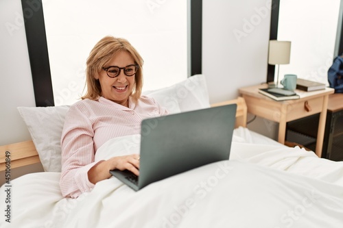Middle age blonde woman smiling happy using laptop lying on the bed at home.