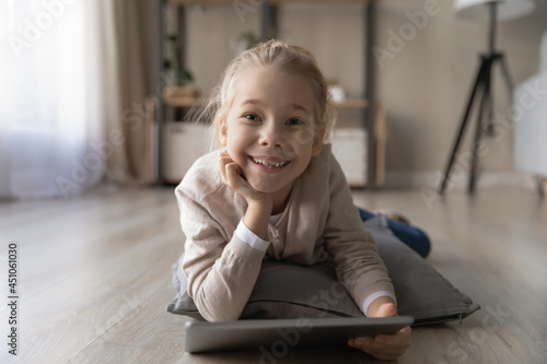 Portrait of happy little teen Caucasian girl child lie relax on floor at home have fun using tablet. Smiling small kid rest talk speak on video call, engaged in webcam virtual event on pad online.