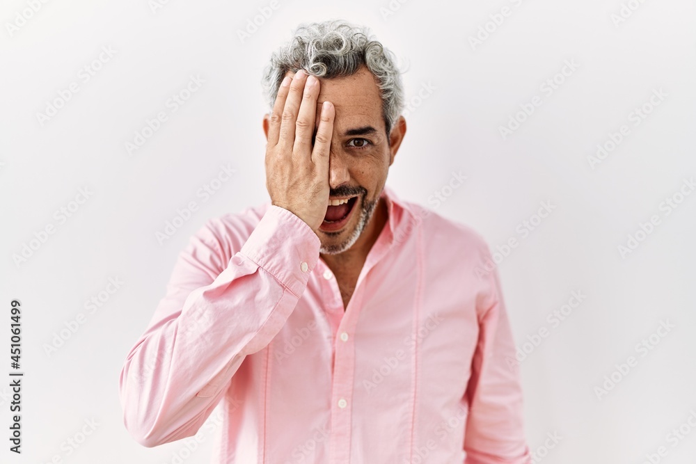 Middle age hispanic man standing over isolated background covering one eye with hand, confident smile on face and surprise emotion. Middle age hispanic man standing over isolated background covering one eye with hand, confident smile on face and surprise emotion.