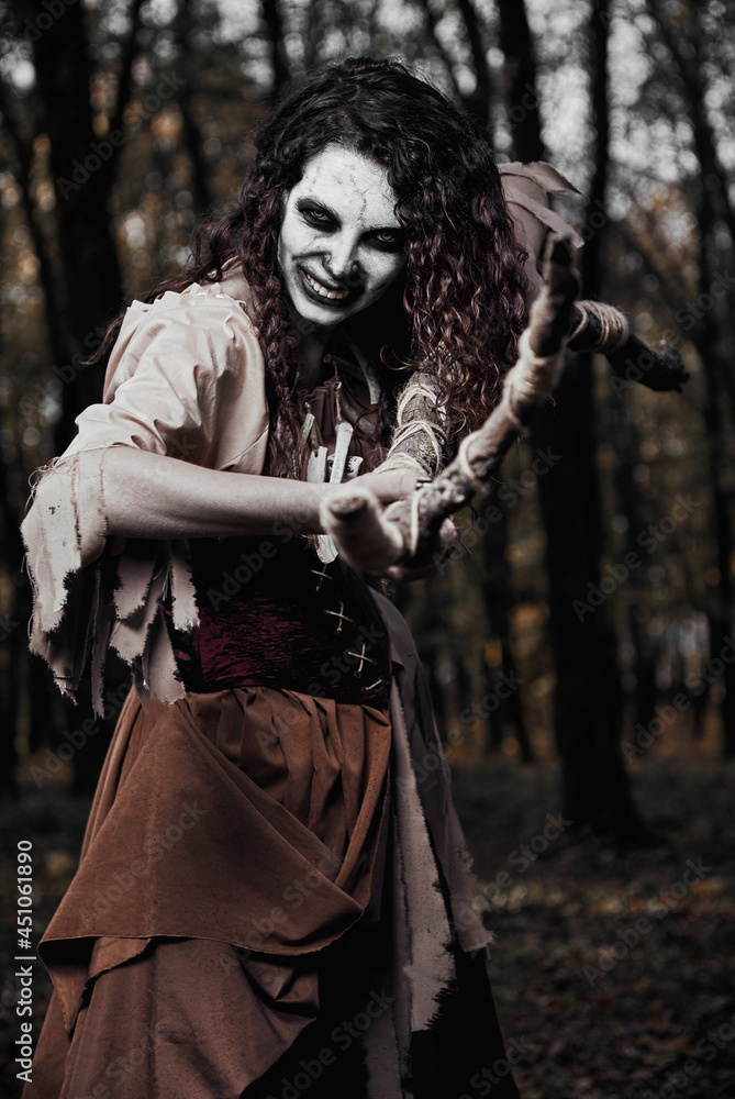 Halloween theme: terrible crazy voodoo witch killing by staff. Portrait of evil sorceress in dark forest. Zombie woman (undead)