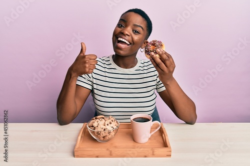 Young african american woman eating breakfast holding chocolate donut smiling happy and positive  thumb up doing excellent and approval sign