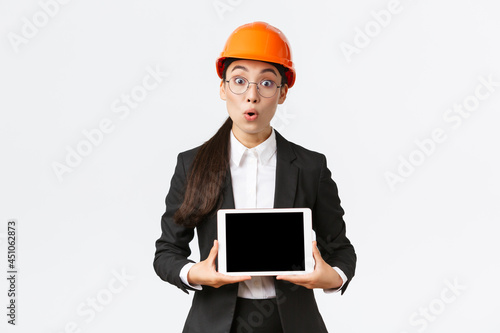 Impressed asian female construction engineer, businesswoman in safety helmet and suit showing chart, introduce digram at digial tablet screen, standing amazed over white background photo