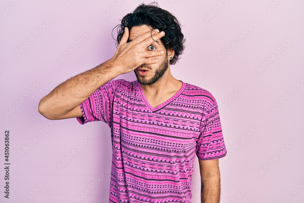 Handsome hispanic man wearing casual pink t shirt peeking in shock covering face and eyes with hand, looking through fingers with embarrassed expression. Handsome hispanic man wearing casual pink t shirt peeking in shock covering face and eyes with hand, looking through fingers with embarrassed expression.