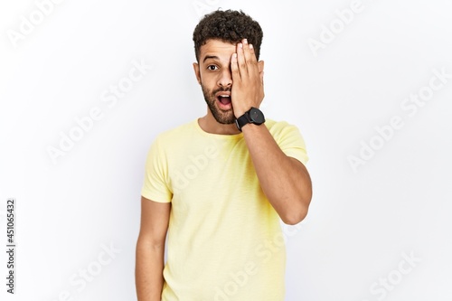 Arab young man standing over isolated background covering one eye with hand, confident smile on face and surprise emotion.