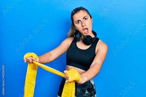 Beautiful hispanic woman training arm resistance with elastic arm band in shock face, looking skeptical and sarcastic, surprised with open mouth