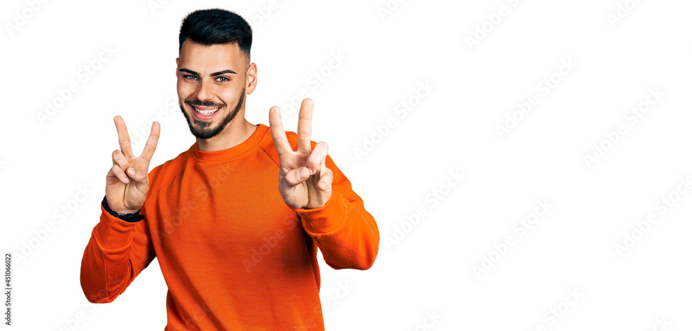 Young hispanic man with beard wearing casual orange sweater smiling looking to the camera showing fingers doing victory sign. number two. Young hispanic man with beard wearing casual orange sweater smiling looking to the camera showing fingers doing victory sign. number two.