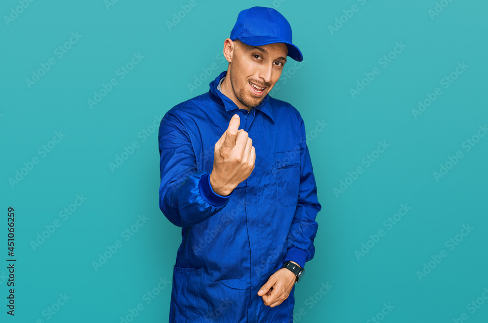 Bald man with beard wearing builder jumpsuit uniform beckoning come here gesture with hand inviting welcoming happy and smiling