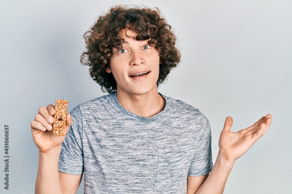 Handsome young man eating protein bar as healthy energy snack celebrating achievement with happy smile and winner expression with raised hand Handsome young man eating protein bar as healthy energy snack celebrating achievement with happy smile and winner expression with raised hand