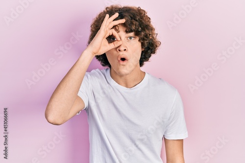 Handsome young man wearing casual white t shirt doing ok gesture shocked with surprised face, eye looking through fingers. unbelieving expression.