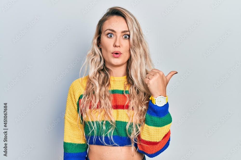 Beautiful young blonde woman wearing colored sweater surprised pointing with hand finger to the side, open mouth amazed expression. Beautiful young blonde woman wearing colored sweater surprised pointing with hand finger to the side, open mouth amazed expression.