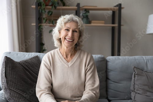 Portrait of smiling old Caucasian 60s woman sit rest on sofa in cozy living room feel optimistic. Happy middle-aged 50s female renter or grandmother relax on couch at home talk on webcam call.