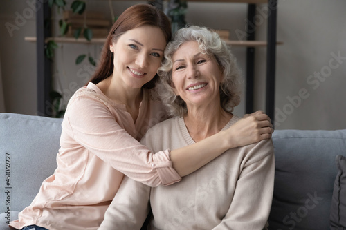 Portrait of two generations of Caucasian women relax on couch at home hugging cuddling together. Smiling adult grownup daughter and older mature mother embrace show love and care in family relations.