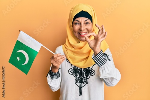 Middle age hispanic woman wearing hijab holding pakistan flag doing ok sign with fingers, smiling friendly gesturing excellent symbol
