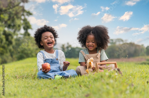 Two African dark skinned children boy and girl sitting on field grass and eating fruits together in parks and outdoors.