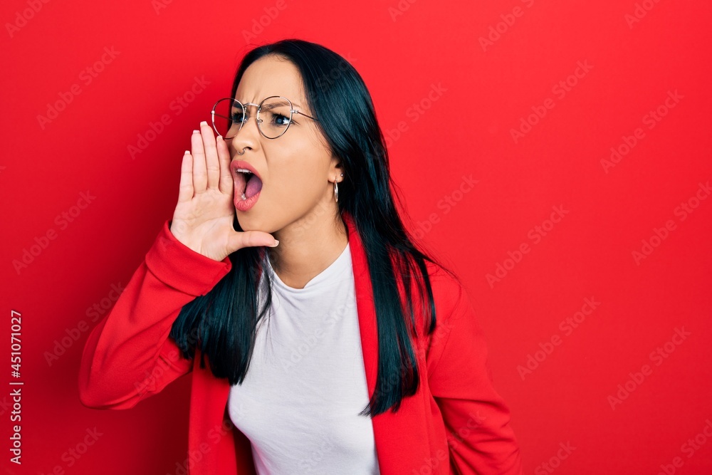 Fototapeta premium Beautiful hispanic woman with nose piercing wearing casual look and glasses shouting and screaming loud to side with hand on mouth. communication concept.