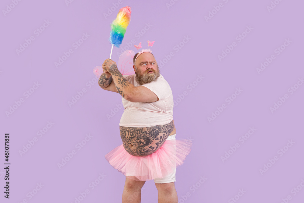 Bellicose mature actor man with overweight in carnival fairy suit with pink wings holds dust brush on purple background in studio