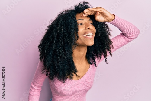 African american woman with afro hair wearing casual pink shirt very happy and smiling looking far away with hand over head. searching concept.