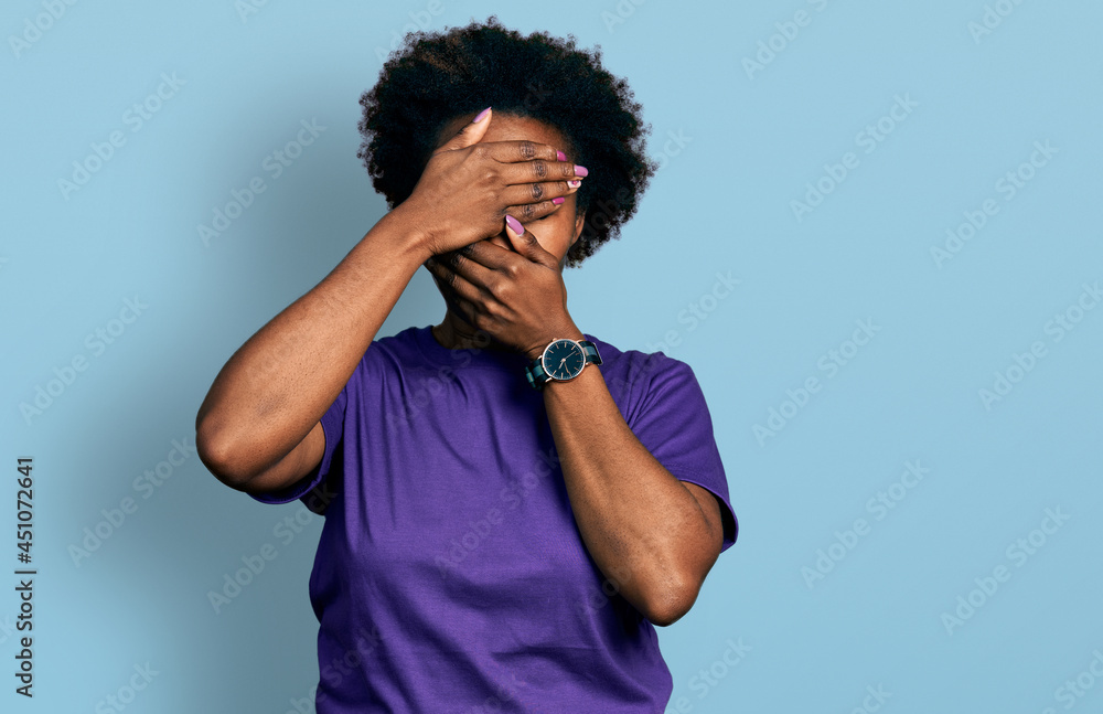 African american woman with afro hair wearing casual purple t shirt covering eyes and mouth with hands, surprised and shocked. hiding emotion African american woman with afro hair wearing casual purple t shirt covering eyes and mouth with hands, surprised and shocked. hiding emotion