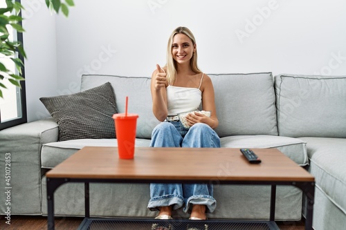 Young blonde woman eating popcorn sitting on the sofa smiling happy and positive, thumb up doing excellent and approval sign