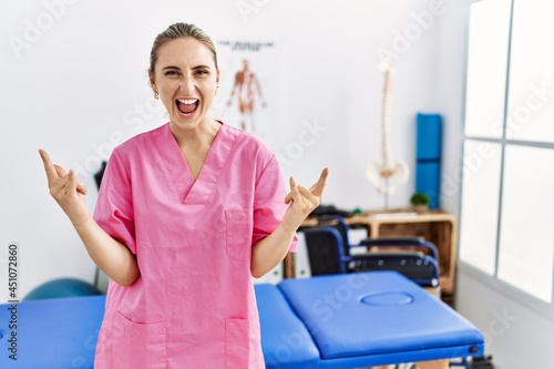 Young blonde woman working at pain recovery clinic shouting with crazy expression doing rock symbol with hands up. music star. heavy concept.