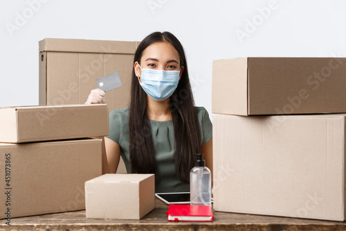 Small business owners, e-commerce and covid-19 preventing virus concept. Asian businesswoman with internet shop wear medical mask, gloves and recommend use credit cards for paying during coronavirus photo