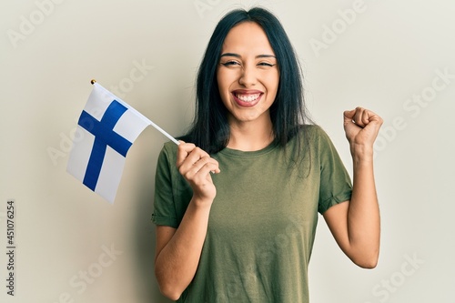 Young hispanic girl holding finland flag screaming proud, celebrating victory and success very excited with raised arm