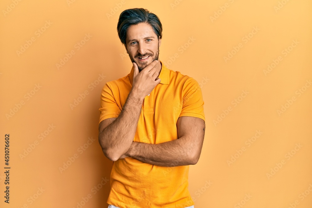 Fototapeta premium Young hispanic man wearing casual yellow t shirt looking confident at the camera smiling with crossed arms and hand raised on chin. thinking positive.