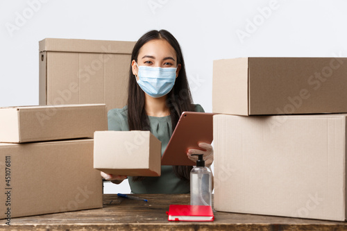 Small business owners, e-commerce and covid-19 preventing virus concept. Asian businesswoman in medical mask, gloves using hand sanitizer while giving out boxes with delivery orders photo