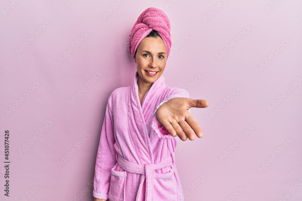 Young blonde woman wearing shower towel cap and bathrobe smiling cheerful offering palm hand giving assistance and acceptance. Young blonde woman wearing shower towel cap and bathrobe smiling cheerful offering palm hand giving assistance and acceptance.