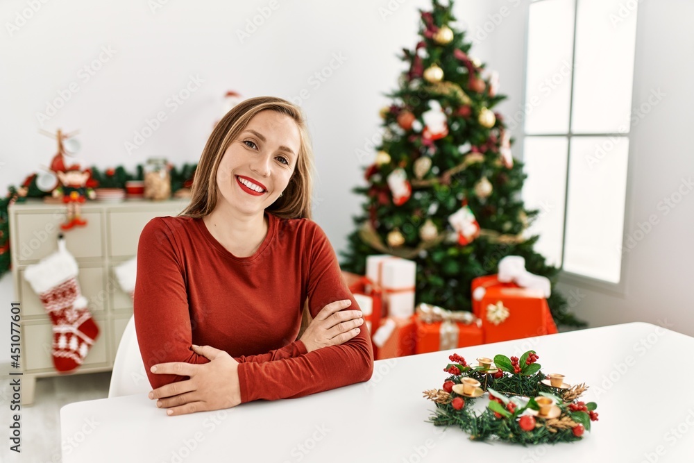 Caucasian young blonde woman sitting on the table by christmas tree happy face smiling with crossed arms looking at the camera. positive person. Caucasian young blonde woman sitting on the table by christmas tree happy face smiling with crossed arms looking at the camera. positive person.