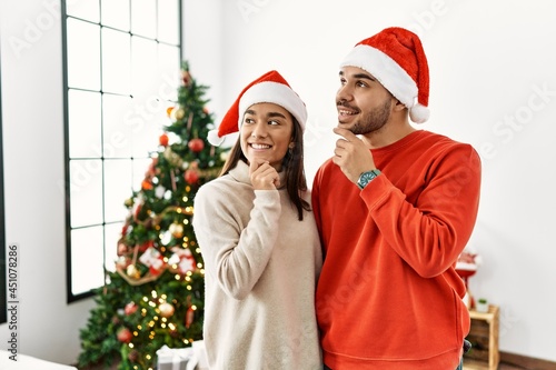 Young hispanic couple standing by christmas tree with hand on chin thinking about question  pensive expression. smiling and thoughtful face. doubt concept.