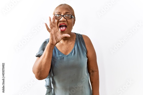 Mature hispanic woman wearing glasses standing over isolated background shouting and screaming loud to side with hand on mouth. communication concept. © Krakenimages.com