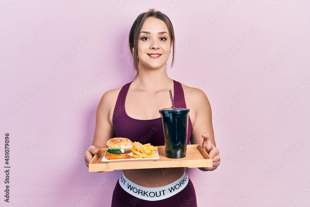 Young hispanic girl wearing sportswear eating a tasty classic burger with fries and soda smiling with a happy and cool smile on face. showing teeth.