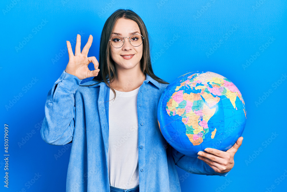 Young hispanic girl holding world ball doing ok sign with fingers, smiling friendly gesturing excellent symbol Young hispanic girl holding world ball doing ok sign with fingers, smiling friendly gesturing excellent symbol