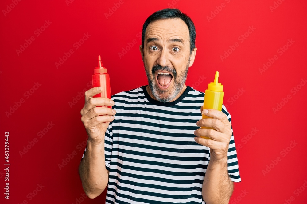 Middle age hispanic man holding ketchup and mustard bottle celebrating crazy and amazed for success with open eyes screaming excited. Middle age hispanic man holding ketchup and mustard bottle celebrating crazy and amazed for success with open eyes screaming excited.