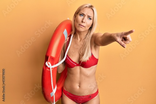 Young blonde woman wearing bikini and holding lifeguard float pointing with finger clueless and confused expression. doubt concept.