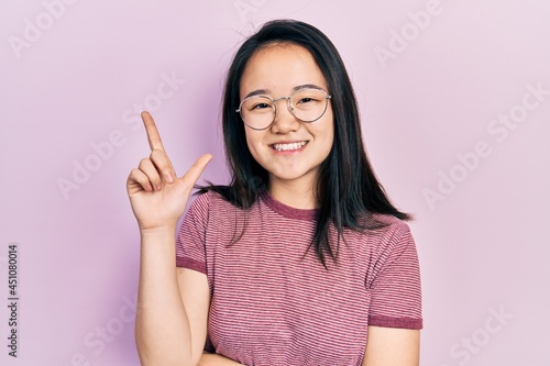 Young chinese girl wearing casual clothes and glasses with a big smile on face  pointing with hand and finger to the side looking at the camera.