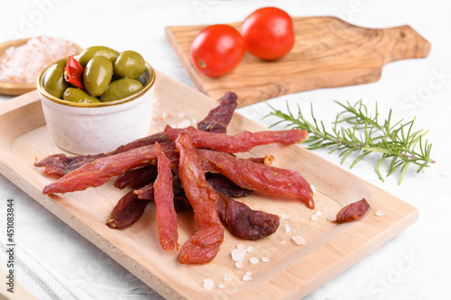 Jerky meat strips with spices, green olives, rosemary on wooden plate on a grey stone background. Tipical Italian food coppiette is a spiced pork meat strips. Snacks for beer photo