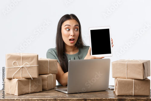 Small business owners, startup and e-commerce concept. Surprised asian businesswoman showing stats on digital tablet screen amazed, packing orders into boxes for shipping, using laptop photo