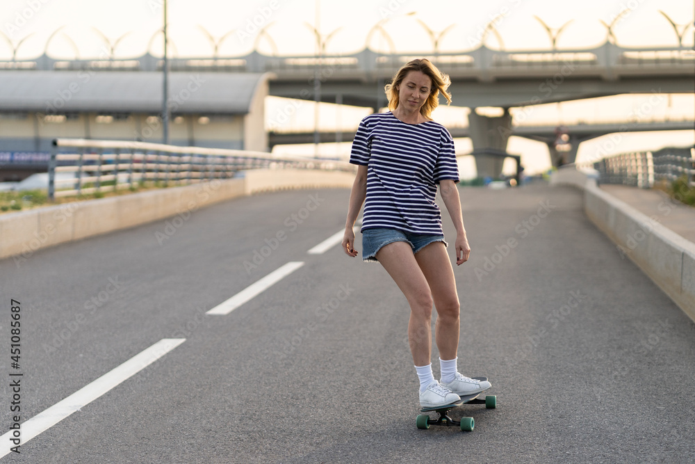 Active woman longboarder of middle age riding longboard on empty road or highway. Casual urban female relaxing on skateboard after work. Lady of 40s skateboarding at sunset. Modern lifestyle concept