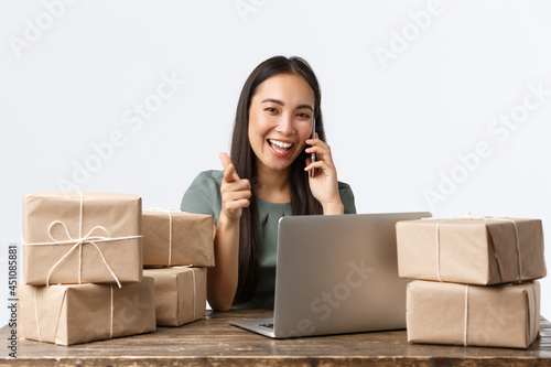 Small business owners, startup and e-commerce concept. Smiling asian businesswoman, online store manager pointing camera, answering client call, taking orders with laptop, packing boxes for shipping photo