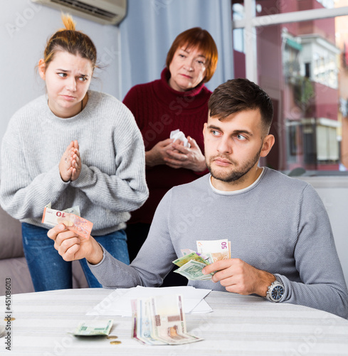 Young woman tearfully asking husband for money while her mother standing behind photo