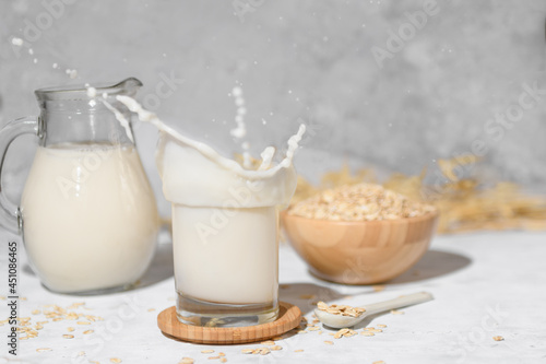 Splash of oat milk in a glass and jug on a grey stone table background and a bowl with oat flake. Jug with vegan non dairy alternative milk.