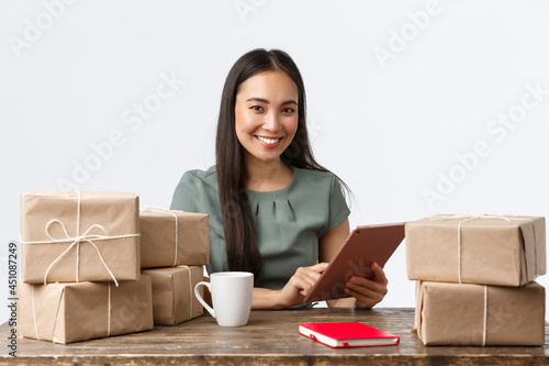 Small business owners, startup and e-commerce concept. Smiling asian businesswoman work from home, packing sold items, wrap boxes with orders for shipping, manage online store via digital tablet photo