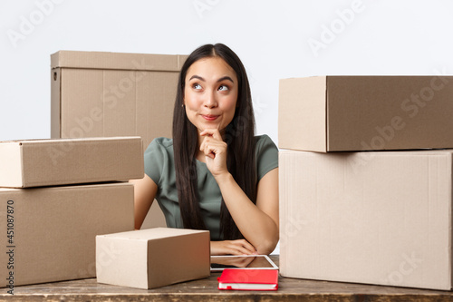 Small business owners, startup and e-commerce concept. Thoughtful creative asian woman open internet store, thinking how make profit, better income, sitting near lots of boxes ready for shipping photo