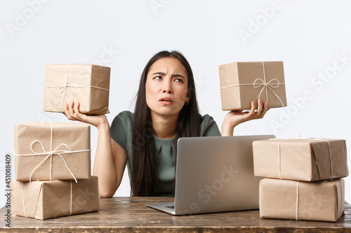 Small business owners, startup and e-commerce concept. Confused asian female entrepreneur working with laptop, packing items for shipping, puzzled as holding boxes, mixed up orders photo