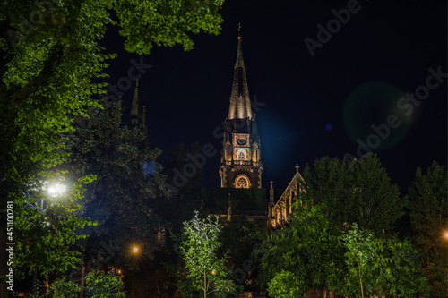 night city gothic cathedral tower long exposure photography foreshortening from below with green tree foliage natural frame foreground and clear star sky background, electricity light and glare effect
