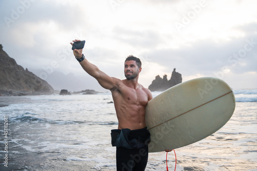 Athletic surfer takes a selfie at the beach, sporty guy holds a surfboard and stands at a tropical beach during sunset - sporty millennial using technology and practicing extreme sport